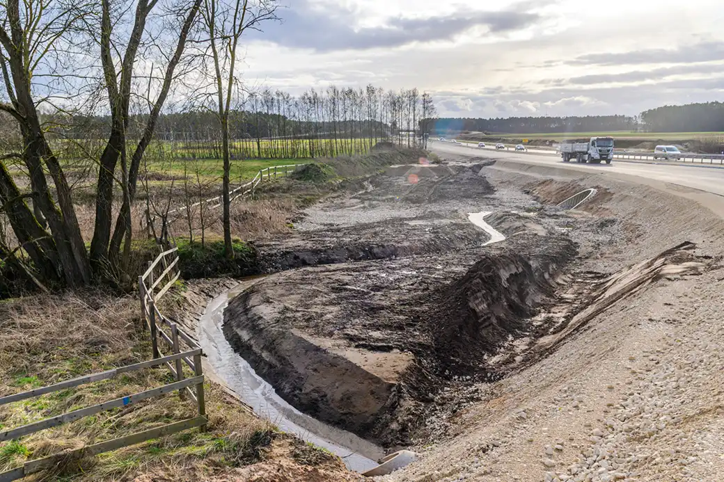 Schlammiger Graben an der Autobahn; kahle Bäume und eingezäunte Felder auf der linken Seite.