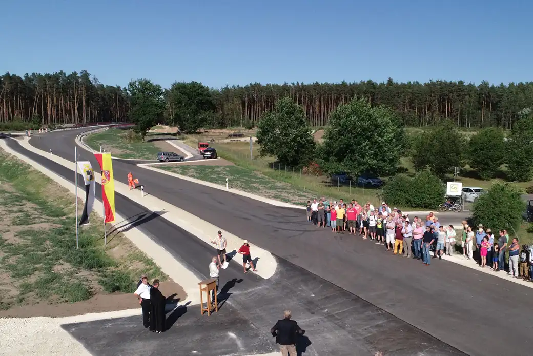 Bei klarem Himmel versammeln sich Menschen mit Fahnen an einer neuen Straße und einem Podium.