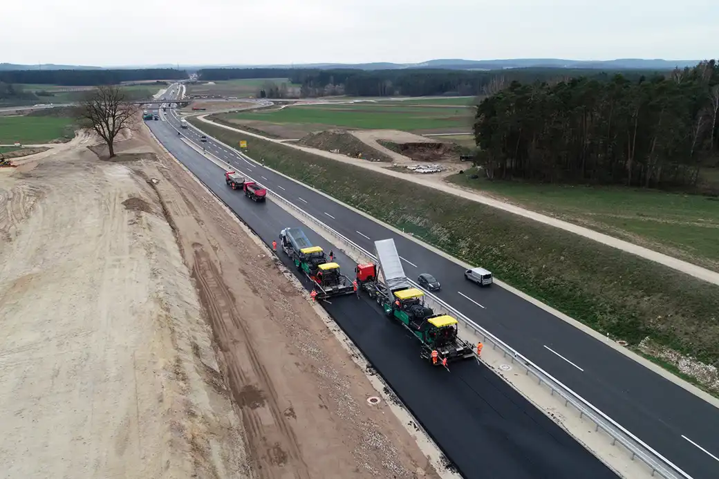 Luftaufnahme von Arbeitern und Fahrzeugen, die eine Autobahn durch Felder und Wälder pflastern.