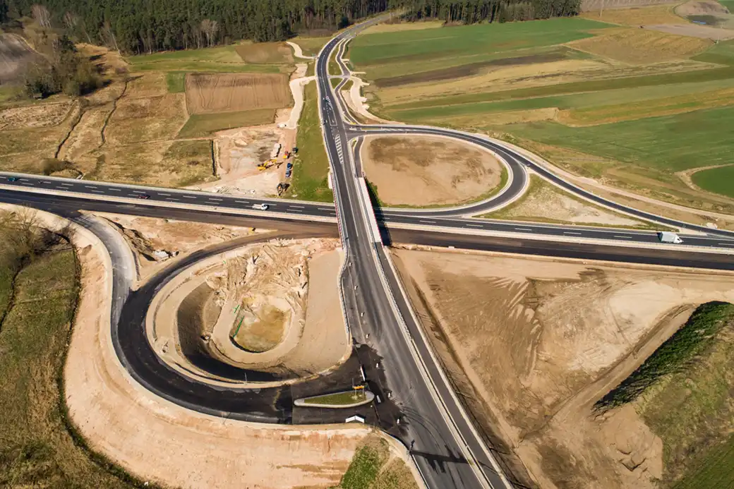 Luftaufnahme: Im Bau befindliches Autobahnkreuz inmitten von Grünflächen und Rampen.