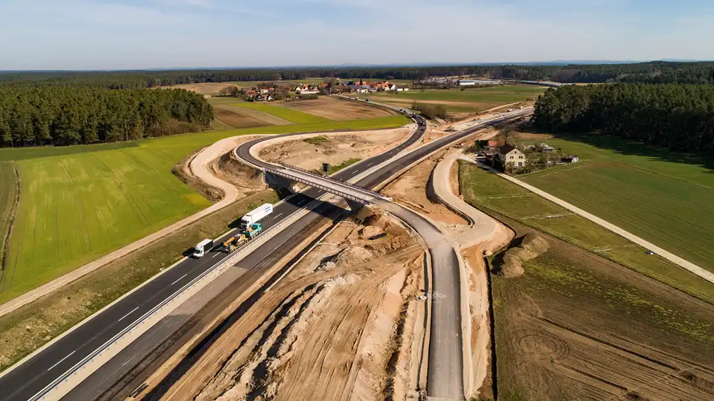 Luftaufnahme: Bau eines Autobahnkreuzes, gepflasterte Straßen, Fahrzeuge, Felder, Wälder.