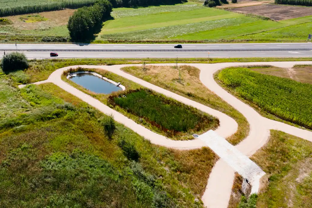 Ein geschwungener Kiesweg führt um den Teich herum, neben der Straße und den grünen Feldern.