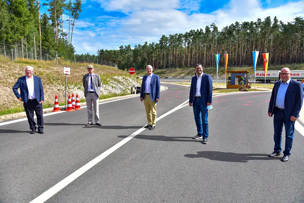 Fünf Geschäftsleute stehen abseits an einer neuen Straße, dahinter Wald und Buden.