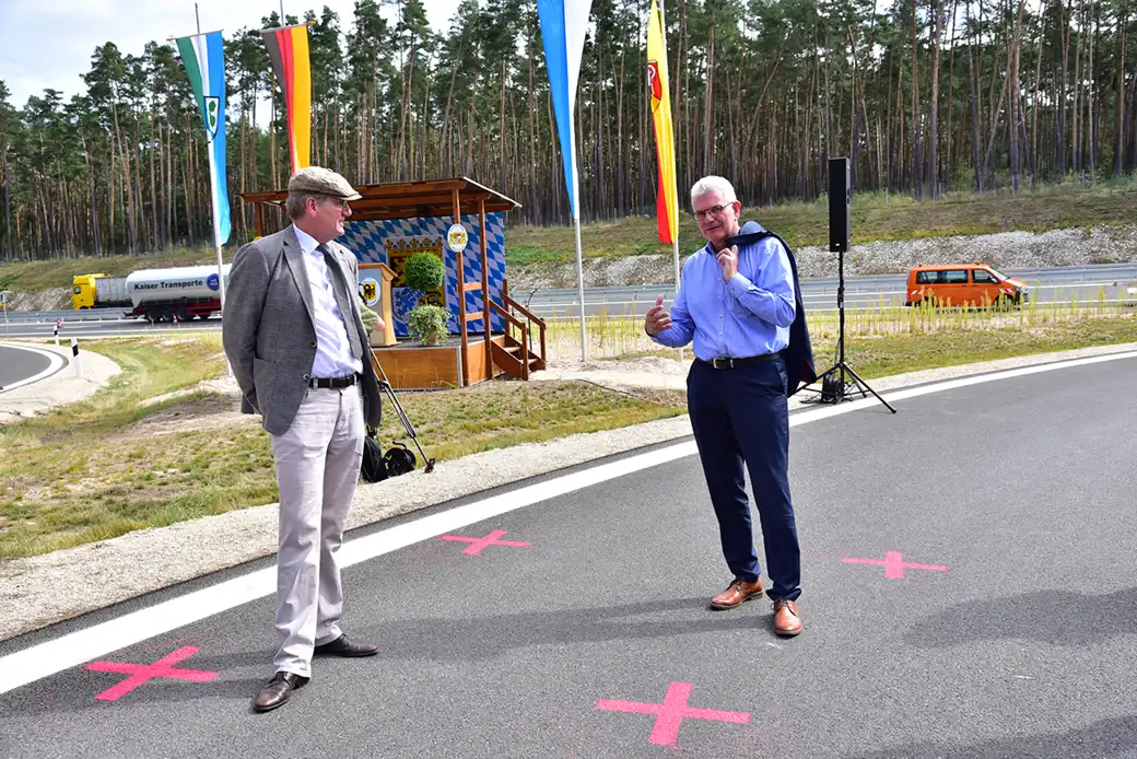 Zwei Männer unterhalten sich auf einer Straße mit rosa X, in der Nähe von Fahnen und einem Lastwagen.