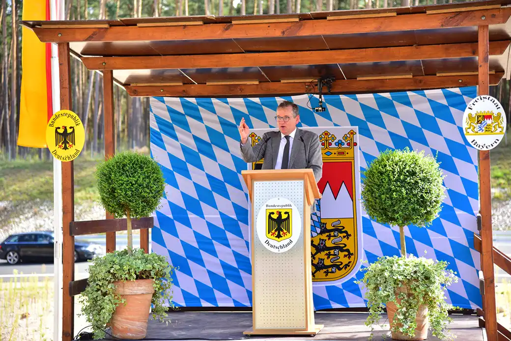 Ein Mann im Anzug spricht auf einem Podium im Freien, das mit bayerischen Motiven geschmückt ist.