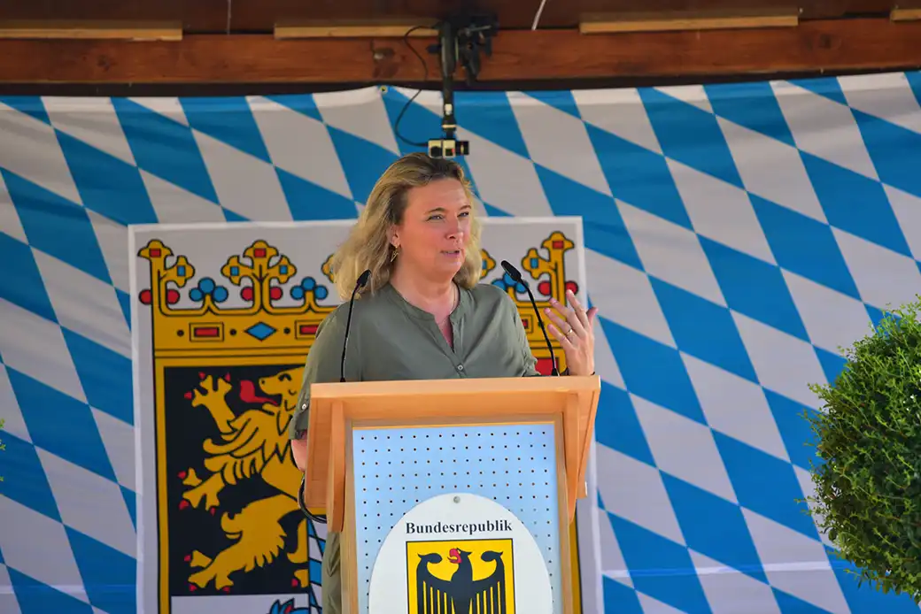 Frau auf Podium mit Aufschrift Bundesrepublik, vor bayerischer Flagge, Mantel.