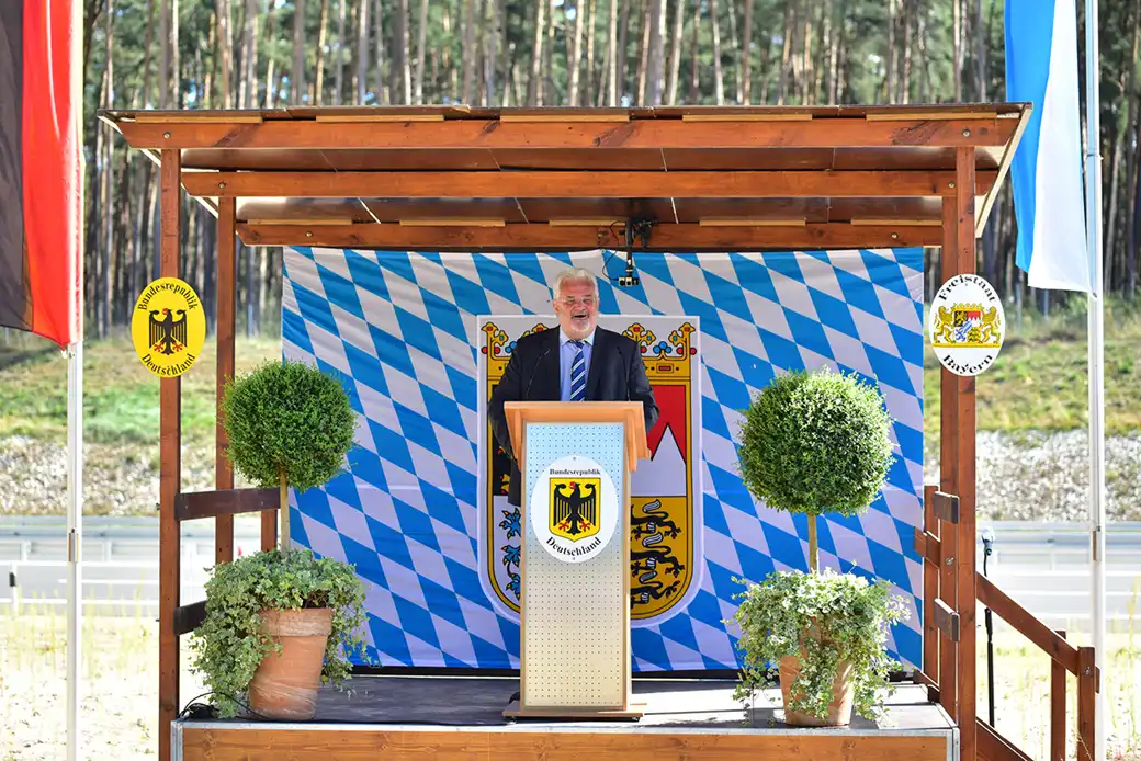 Ein Mann hält eine Rede auf einer Bühne im Freien mit bayerischen Fahnen und Dekorationen.