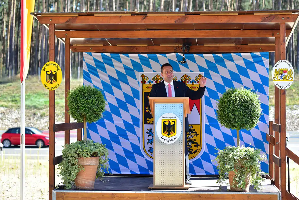 Mann im Anzug spricht am Podium auf einer Außenbühne mit bayerischem Dekor.