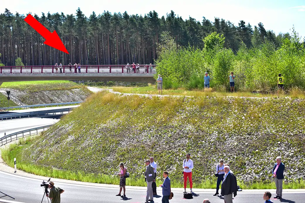 Menschen stehen im Gras und auf der Brücke; der rote Pfeil hebt die Gruppe auf der Brücke hervor.