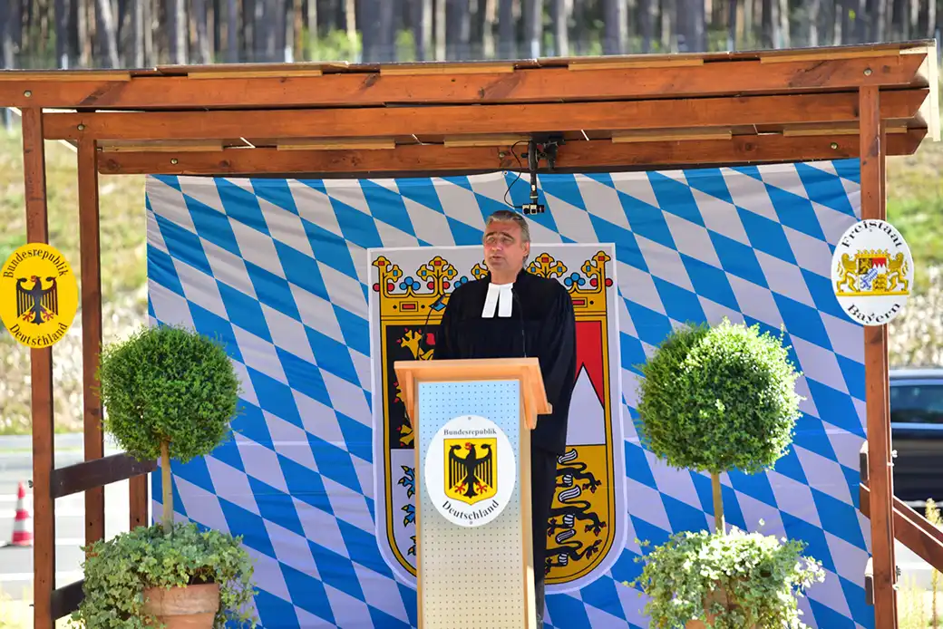 Mann in schwarzer Robe spricht am Podium vor bayerischer Flagge und Emblemen.