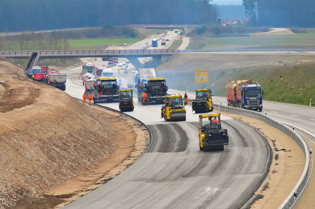 Baufahrzeuge und Arbeiter ebnen eine neue Autobahn inmitten von Bäumen und Wolken.