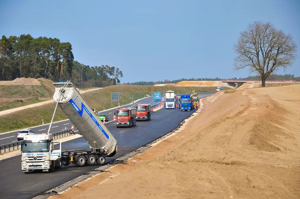Lastwagen und Baufahrzeuge pflastern eine Autobahn; Bäume und Böschungen in der Nähe.