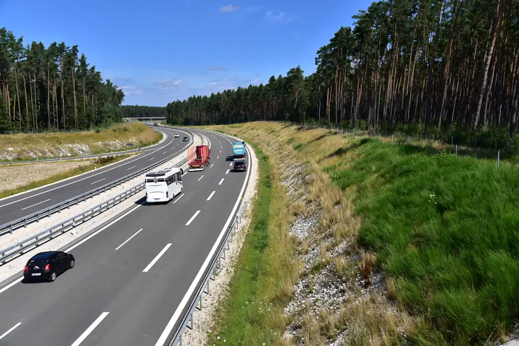 Fahrzeuge fahren auf einer geteilten Autobahn, umgeben von Gras, Wald und Himmel.