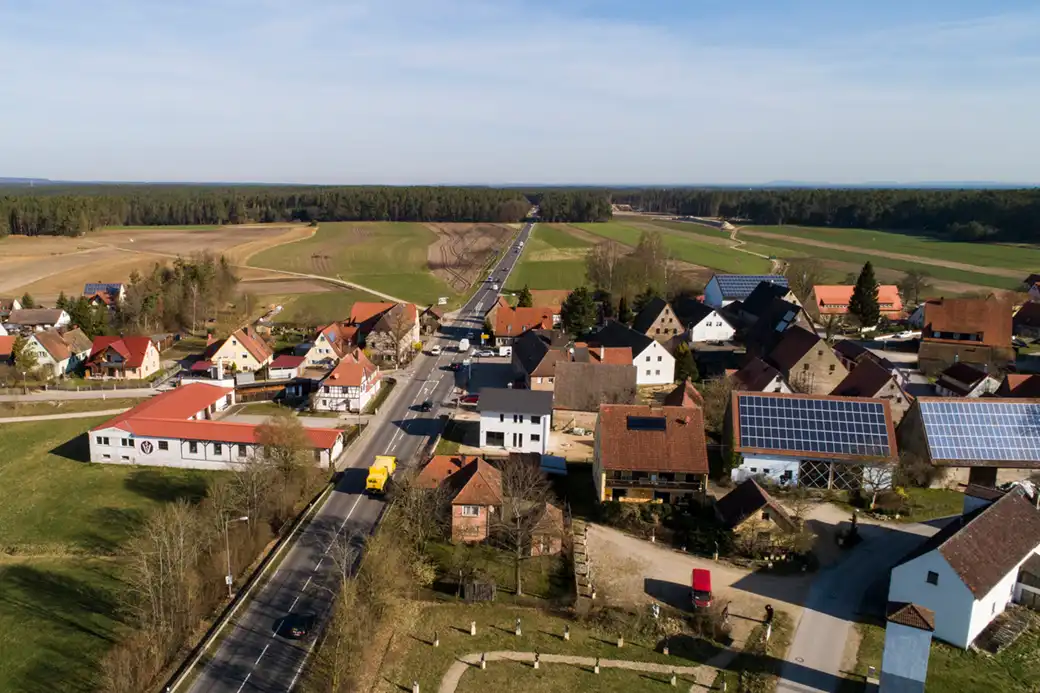 Luftaufnahme eines Dorfes mit Sonnenkollektoren, Feldern und einer zentralen Straße.